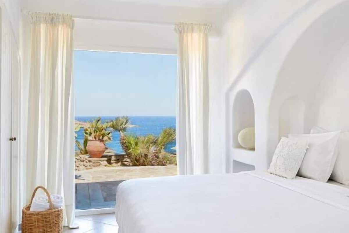 A bedroom with a bed, white curtains, a view of the sea, and a wicker basket.