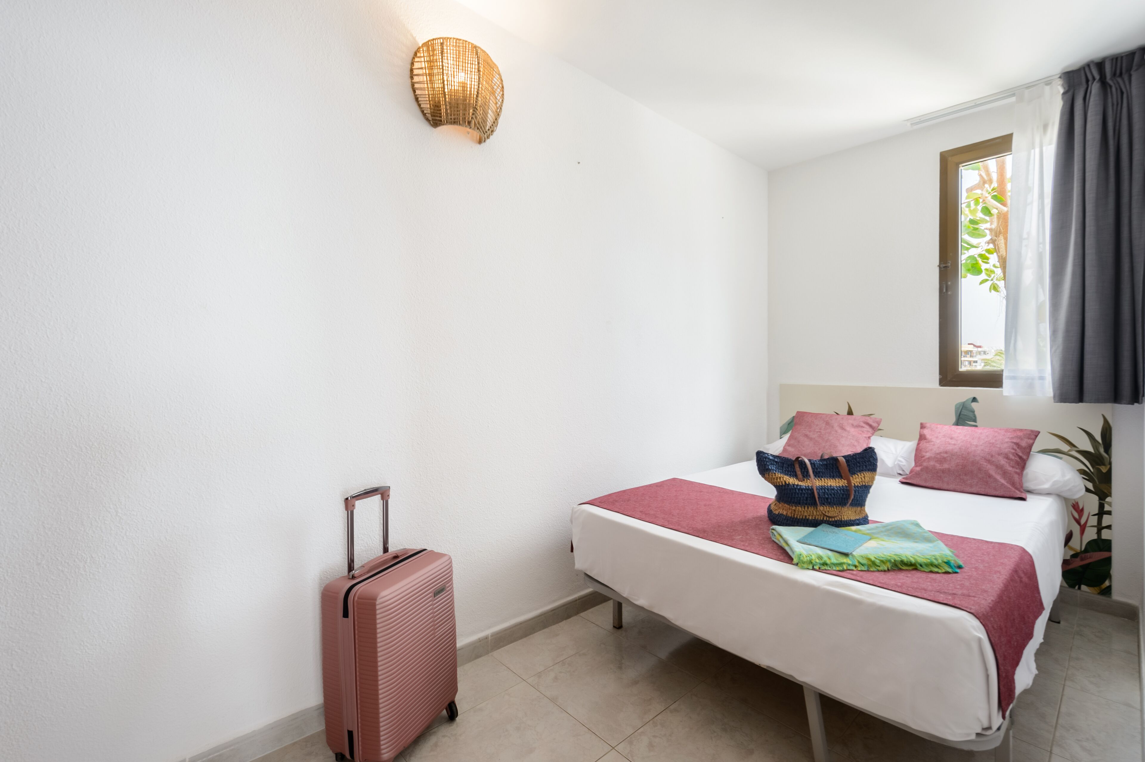 A bedroom with a bed, a suitcase, a wall-mounted light, and a window with curtains.