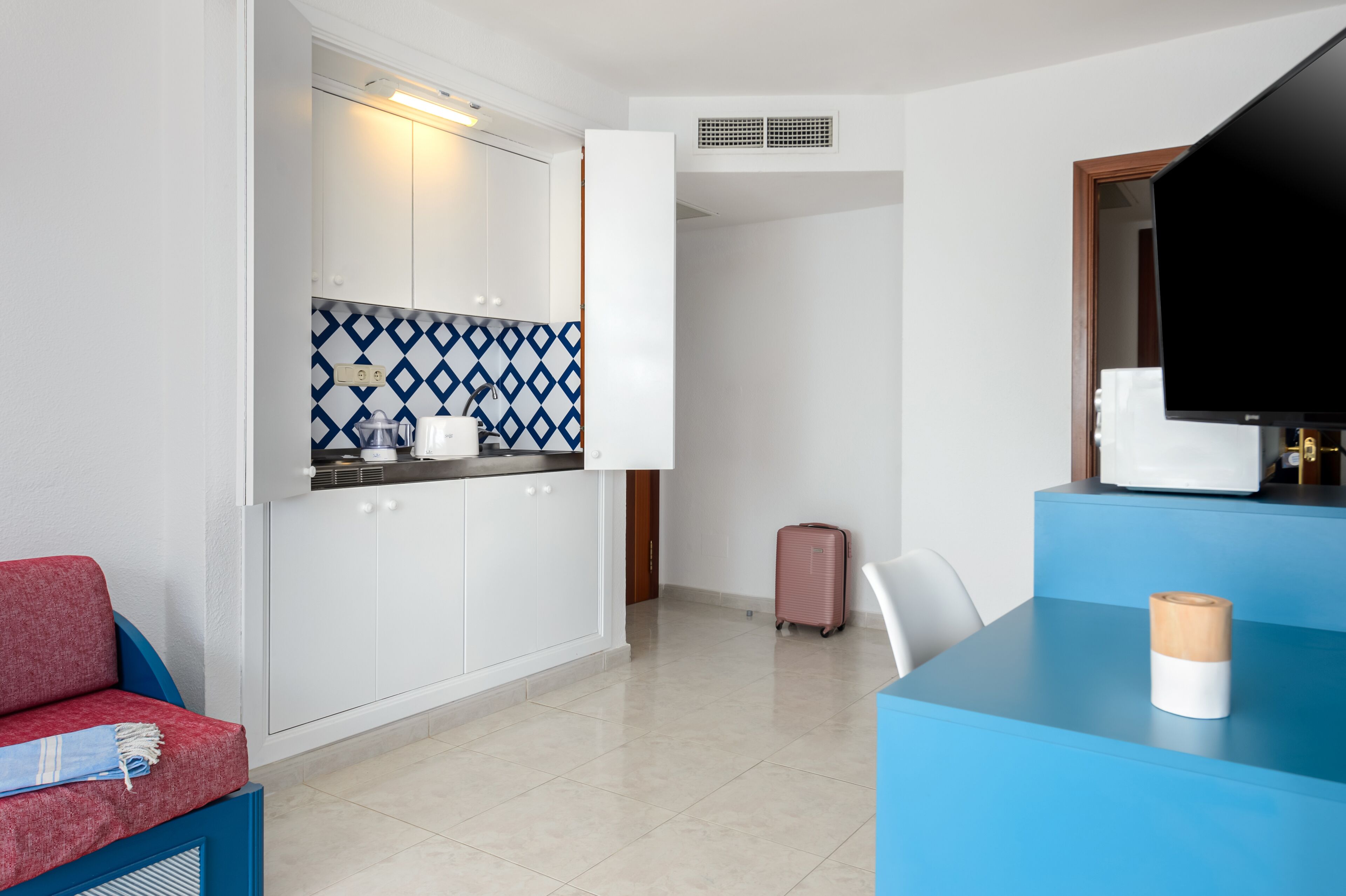 A compact hotel room with a kitchenette, a seating area with a red sofa, a small table with a wooden object, and a television on a stand.
