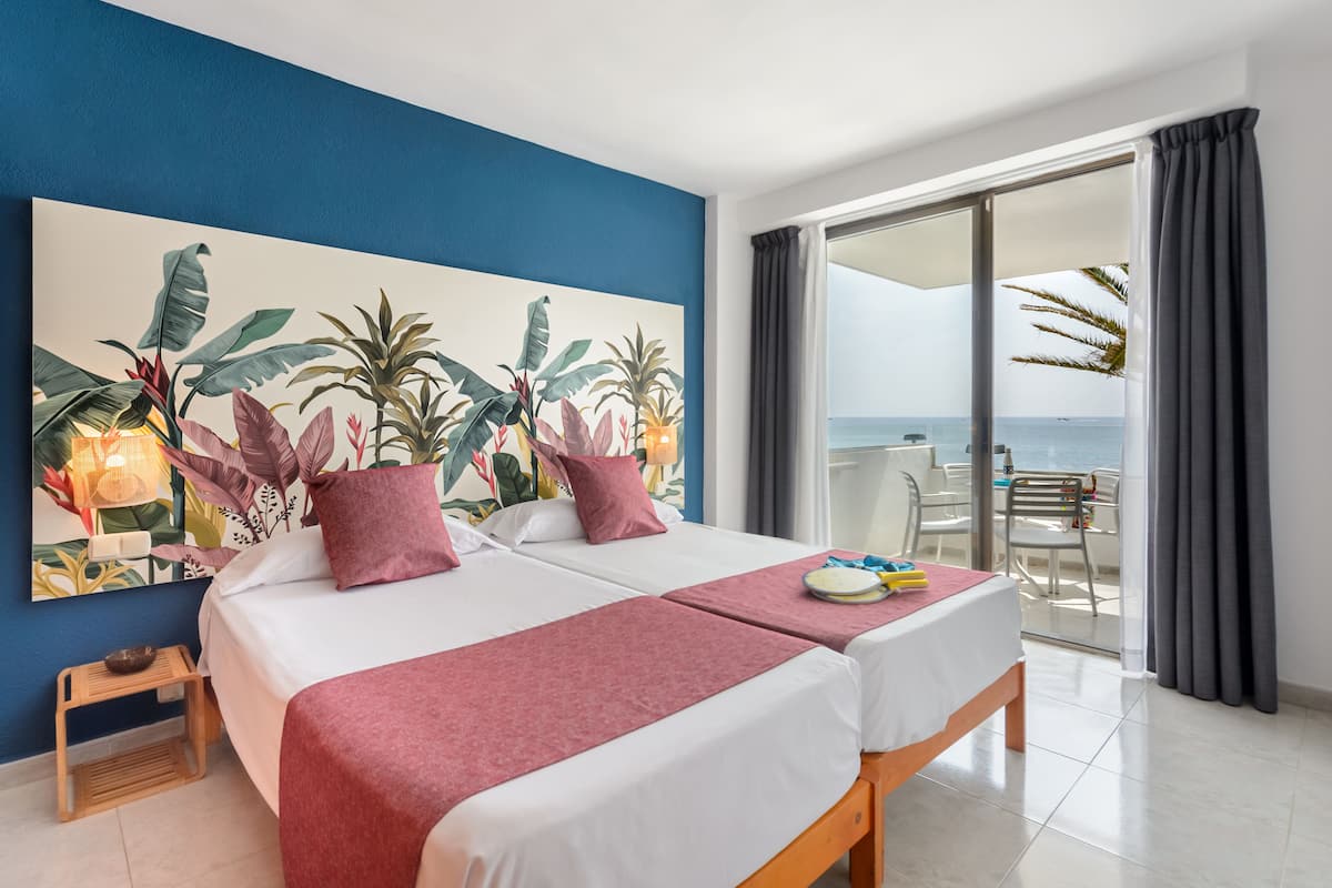 Ein Hotelzimmer mit zwei Betten, einem großen Gemälde im tropischen Stil und einem Balkon mit Meerblick.