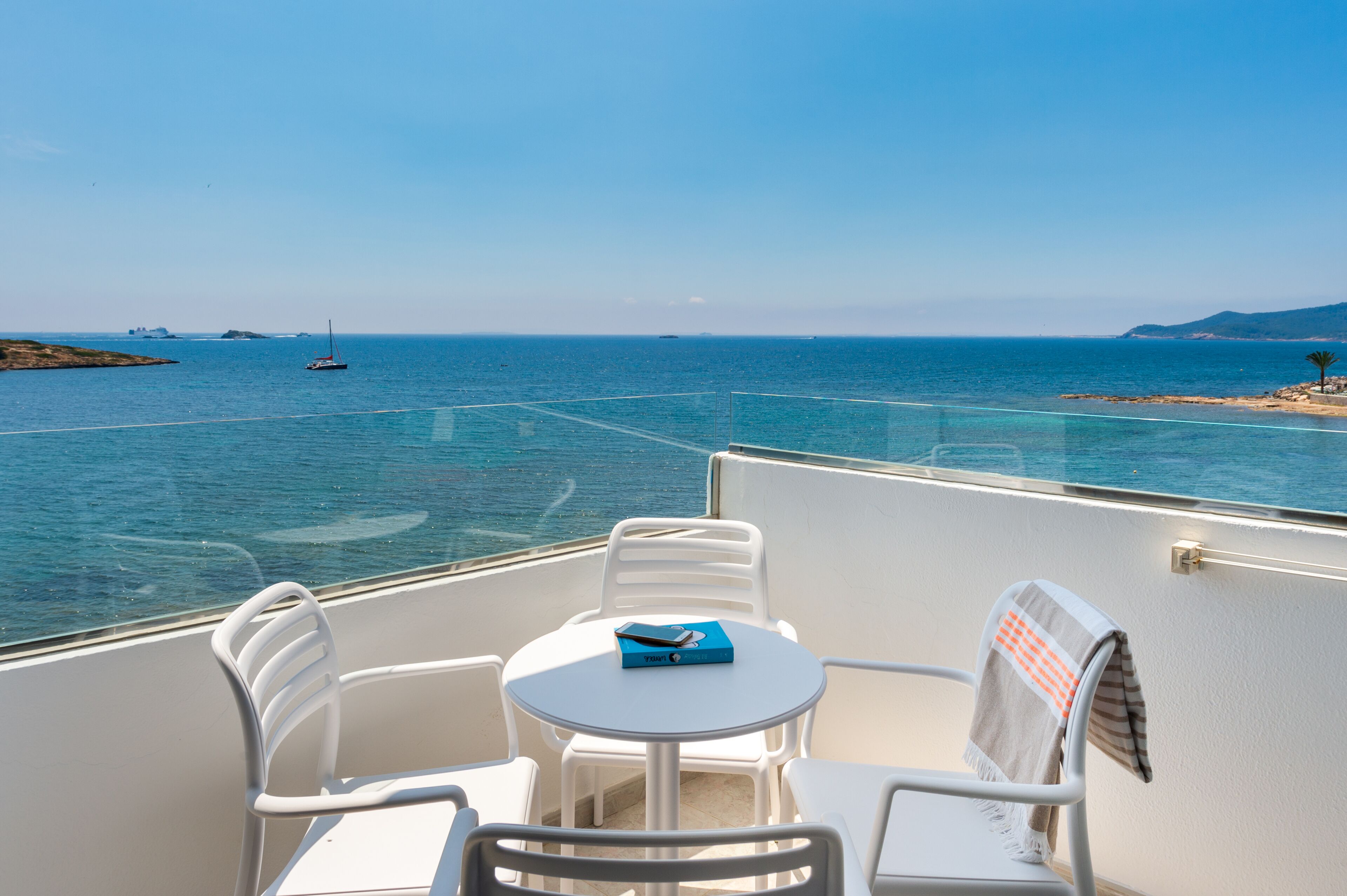 Ein Balkon mit Tisch und Stühlen, Blick auf ein ruhiges Meer mit Segelboot und weiter Küste.