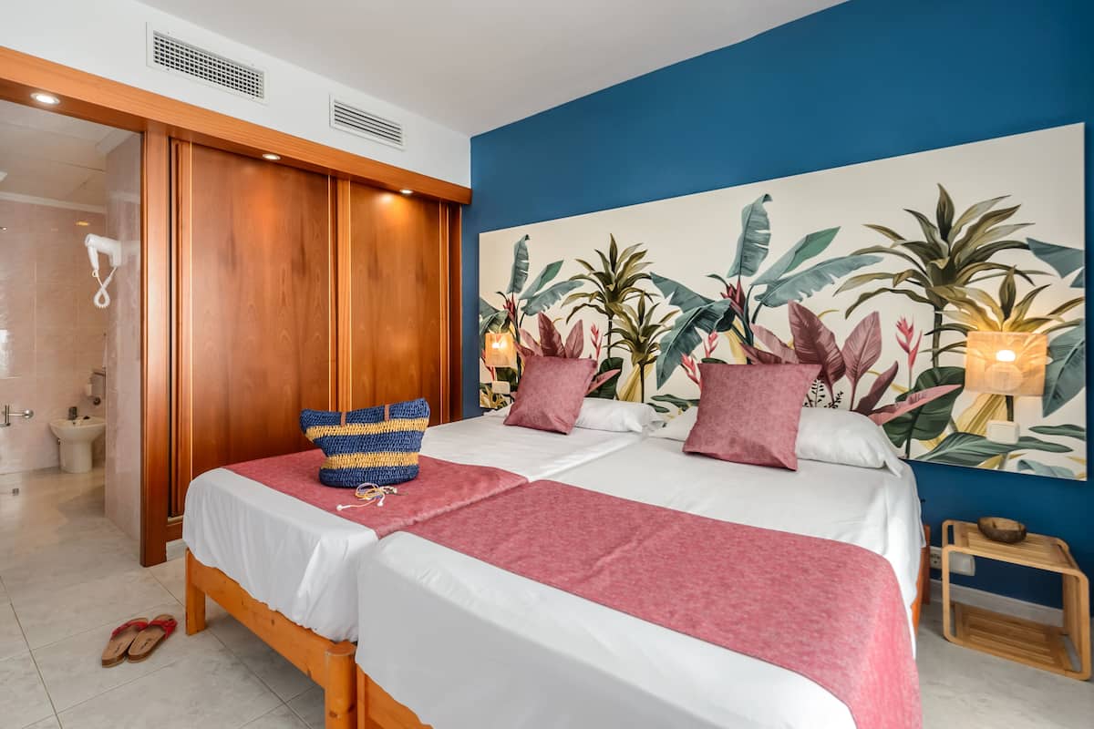 Ein Hotelzimmer mit zwei Betten, einem Holzschrank, einem Badezimmer mit Dusche und einem großen Wandbild im tropischen Stil.