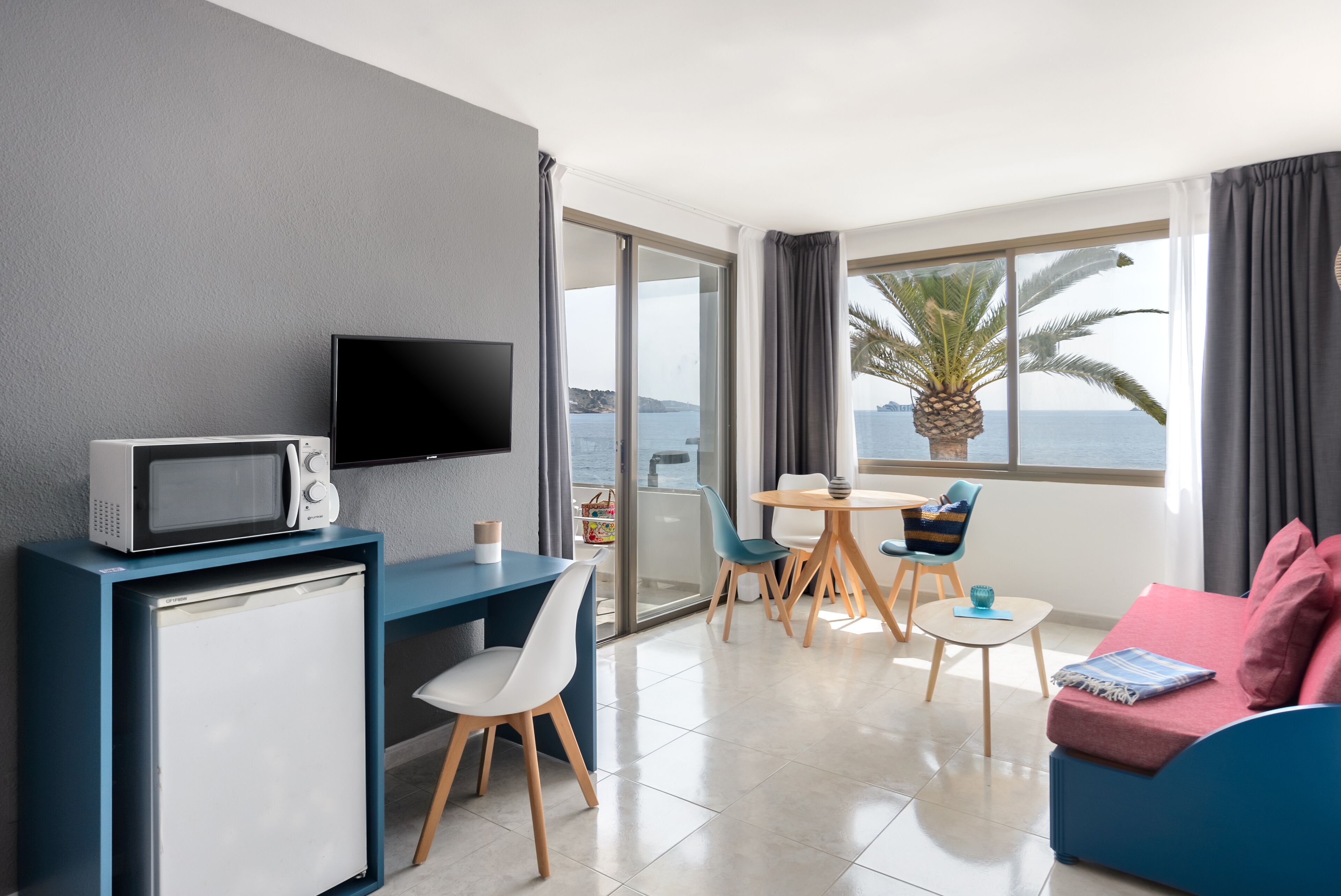A modern hotel room with a flat-screen TV, a compact fridge, a dining area with a table and chairs, a sofa, and a large window with a view of the sea.