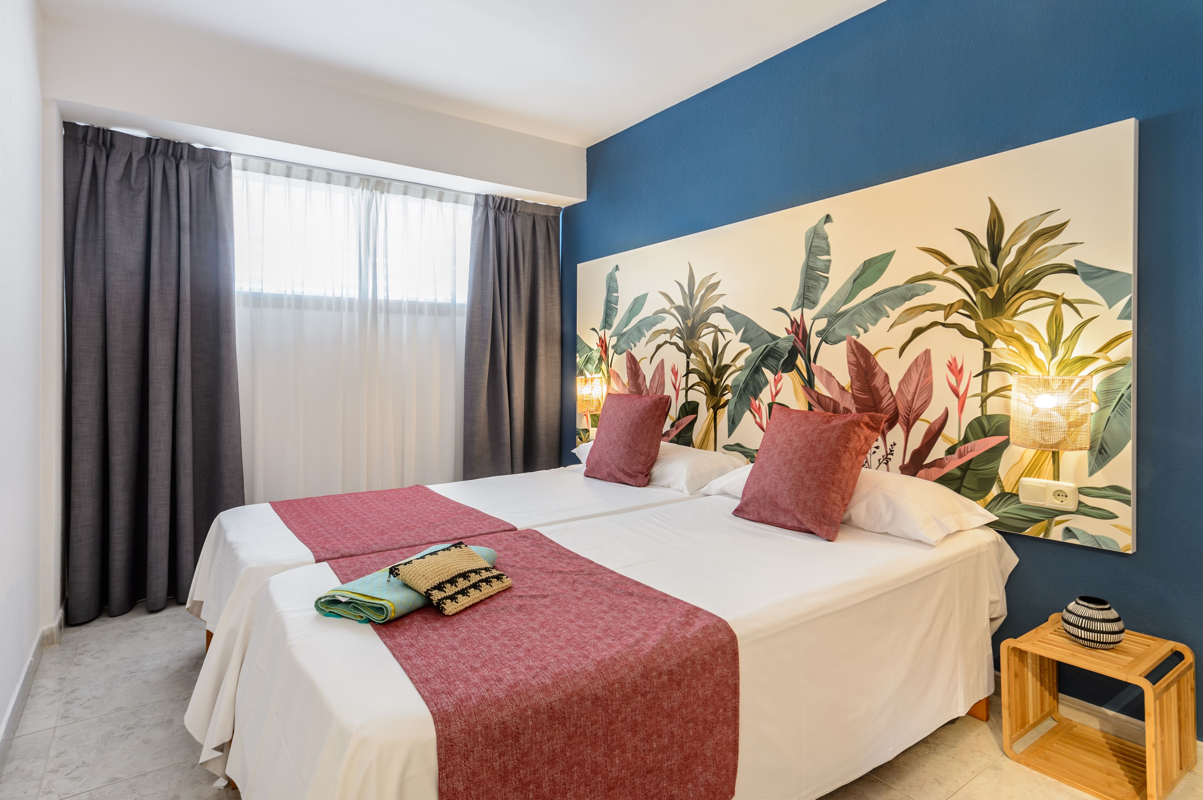 A double bed with a red blanket and pillows, a small wooden side table, a large tropical plant painting on the wall, and a window with grey curtains.