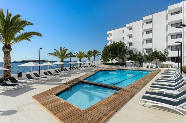 Un espace piscine avec des chaises longues et des parasols, une terrasse en bois et une vue sur la mer.