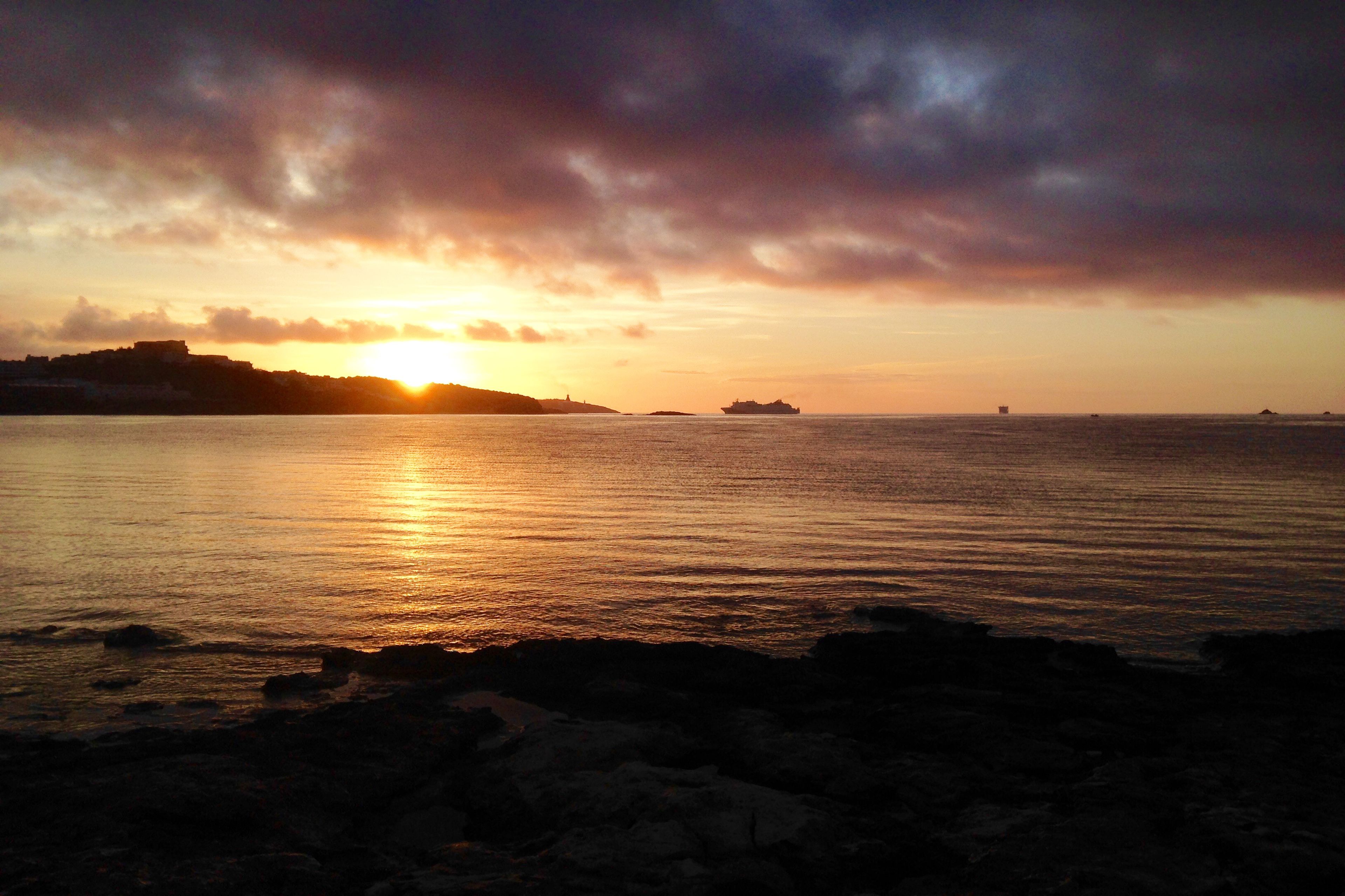 A sunset over a calm sea with a distant shoreline and a ship.