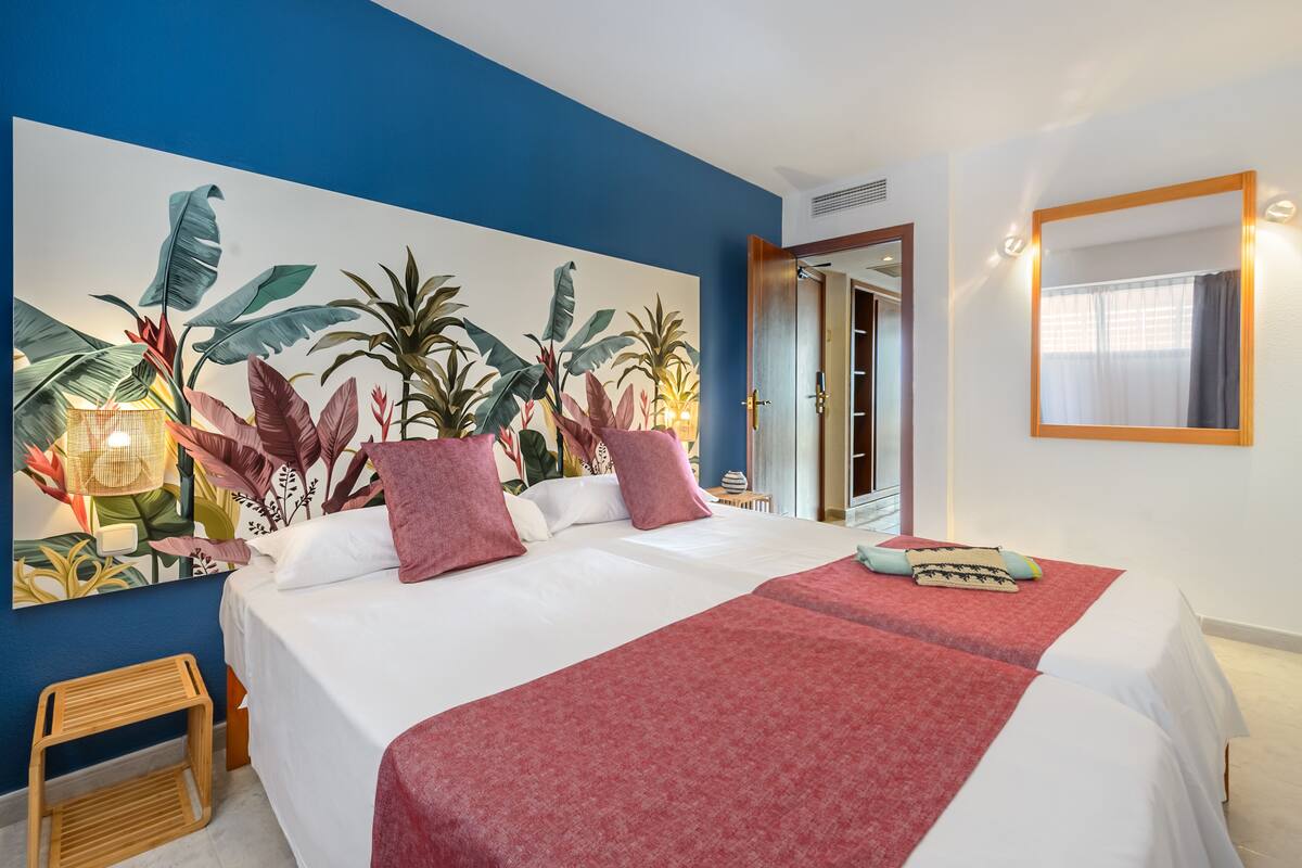 Ein Hotelzimmer mit einem großen Bett, einer roten Bettdecke, einem Wandbild mit tropischen Pflanzen und einem spiegelnden Wandbild mit Holzrahmen.