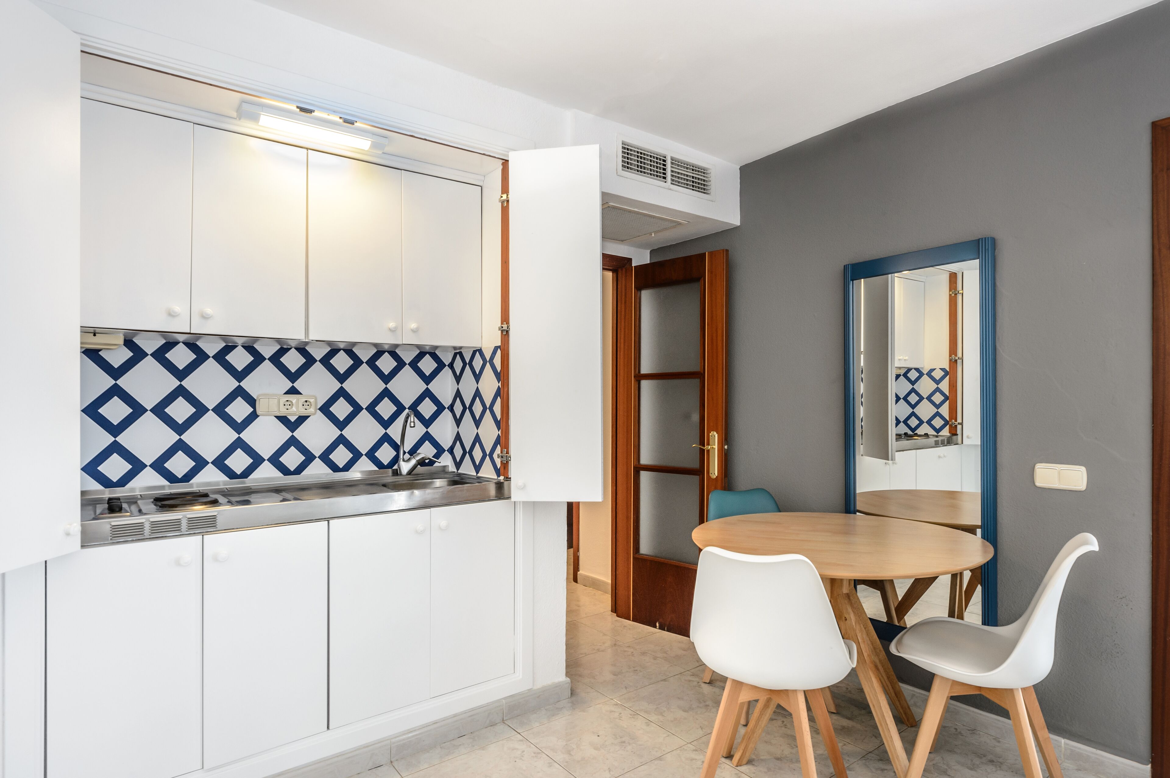 A compact kitchen with white cabinets, a blue and white patterned backsplash, and a wooden dining table with white chairs.