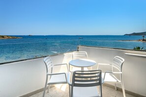 Un balcon avec des chaises et une table en plastique blanc, d’où l’on peut admirer une mer calme, des bateaux et une terre lointaine.