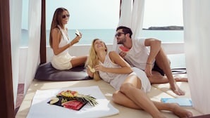 A couple and a woman relaxing on a sunbed by a window overlooking the sea, with a fruit platter on the sunbed. - Apartamentos Vibra Jabeque Soul (Ibiza Town)