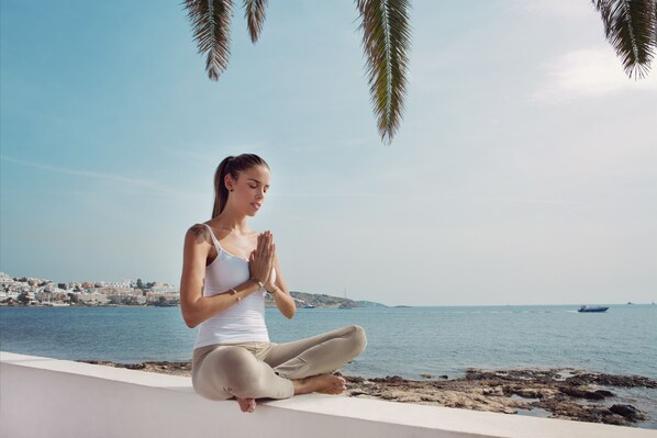 Una persona meditando en un saliente con vista al mar y un paisaje urbano de fondo.