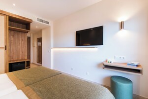 A modern hotel room with a bed, a television mounted on the wall, a small shelf with books and a lamp, and a wooden wardrobe.