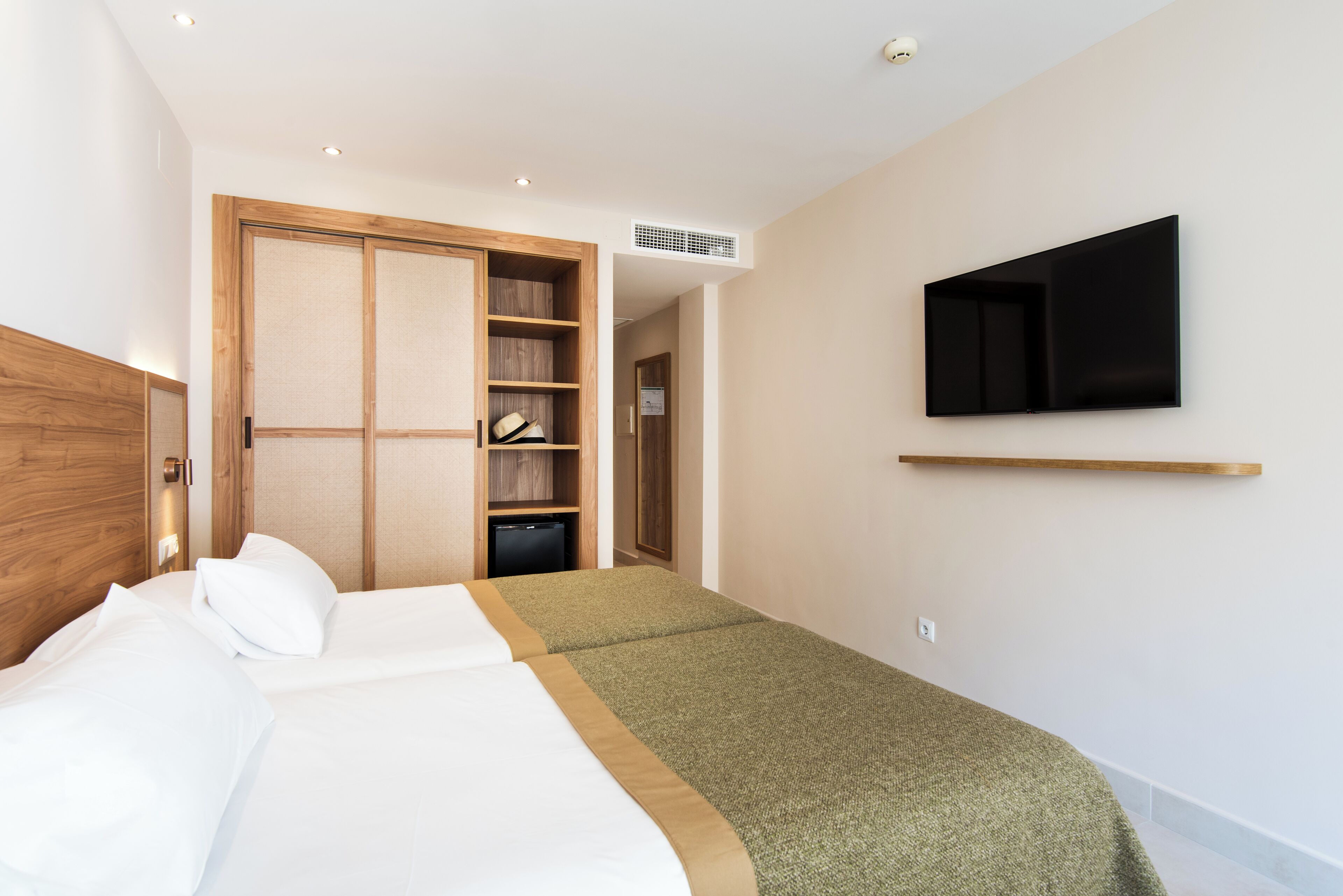 A hotel room with a bed, a TV mounted on the wall, a wooden wardrobe, and a small desk.