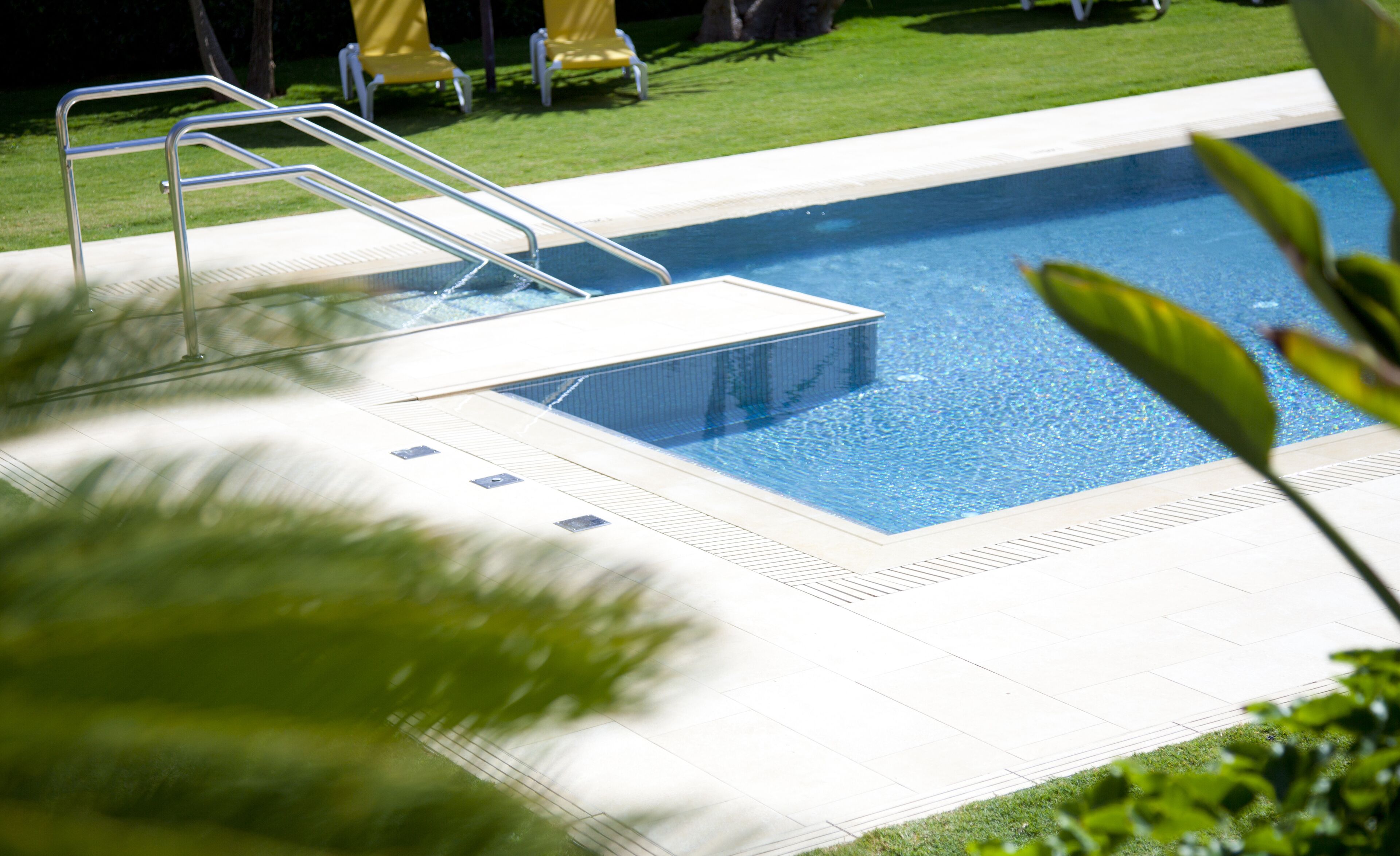 Une piscine avec un escalier en métal, entourée d’une terrasse carrelée et d’une pelouse agrémentée de chaises jaunes.