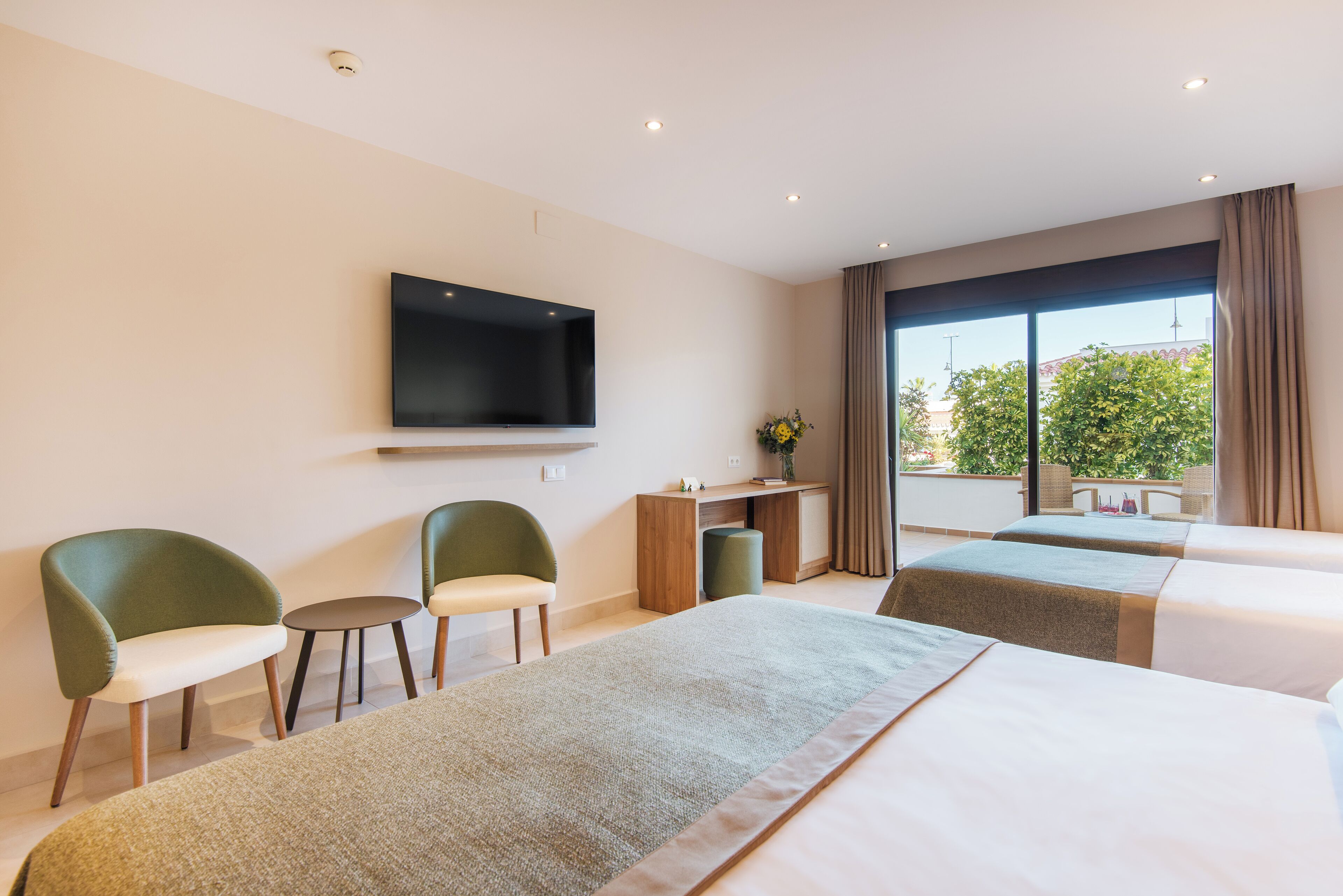 Hotelkamer met een groot bed, twee kleinere bedden, een aan de muur bevestigde televisie, een bureau met stoel en een raam met gordijnen.