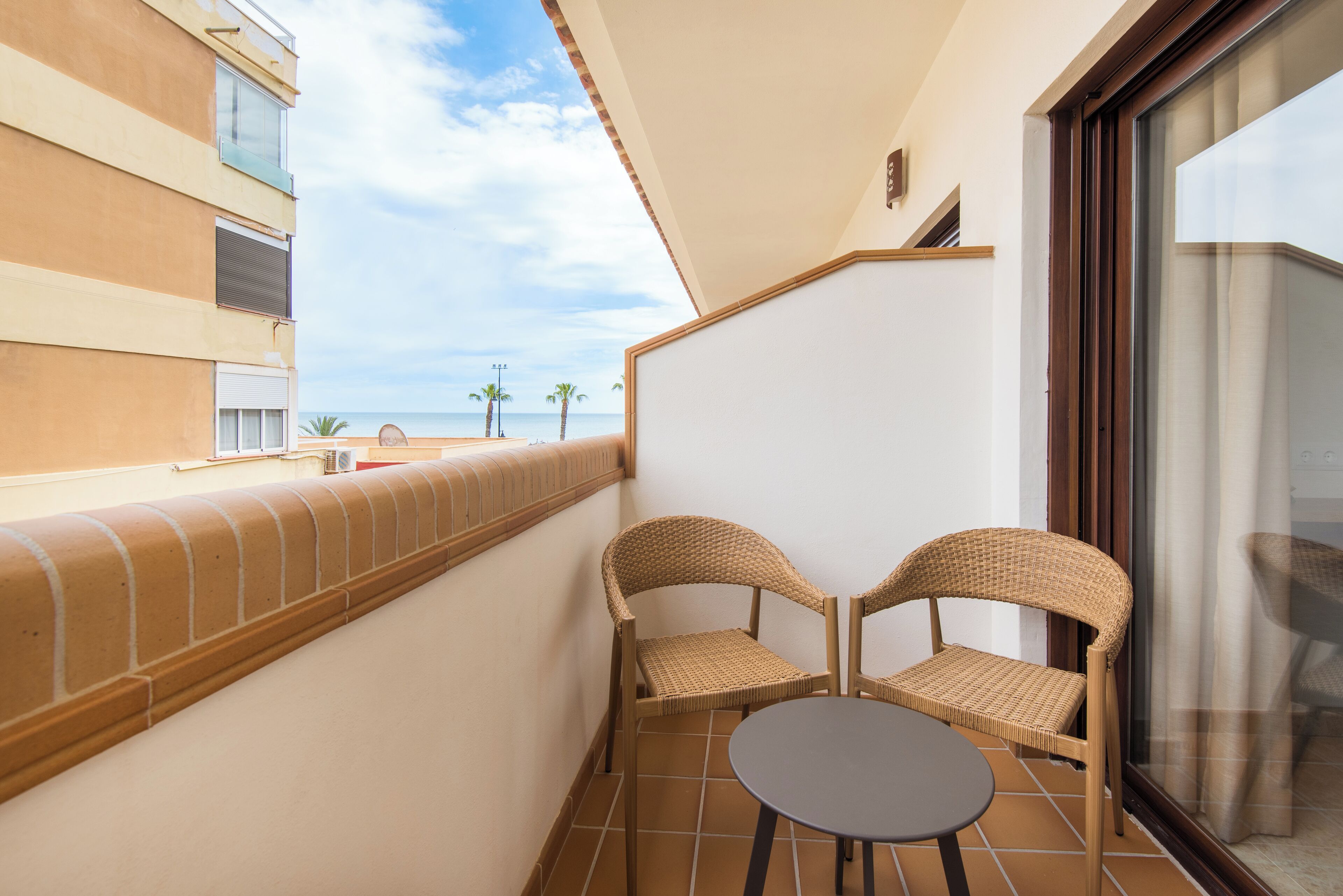 A balcony with two wicker chairs and a small table, overlooking a beach and palm trees.