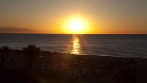 A beach scene with the sun setting over the ocean, palm trees, and beach umbrellas. - Hotel Tarik (Torremolinos)