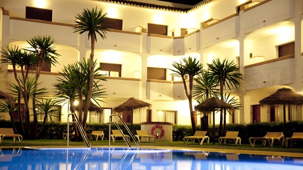 A swimming pool with lounge chairs and palm trees, surrounded by a multi-story building with balconies. - Hotel Tarik (Torremolinos)