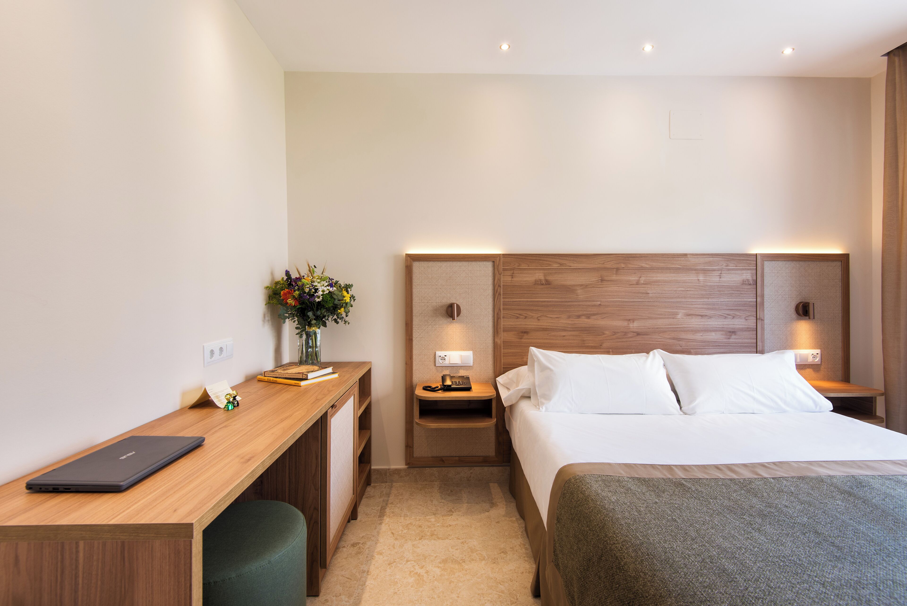 A hotel room with a wooden desk, a bed with white linens, a nightstand with a lamp, and a small vase with flowers.