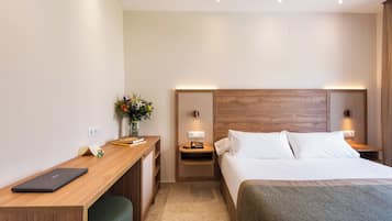 Camera d'albergo con una scrivania in legno, un letto con lenzuola bianche, un comodino con una lampada e un piccolo vaso con fiori.