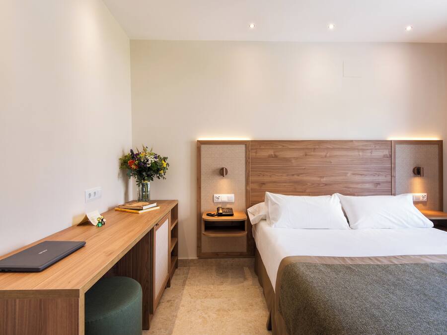 Camera d'albergo con una scrivania in legno, un letto con lenzuola bianche, un comodino con una lampada e un piccolo vaso con fiori.