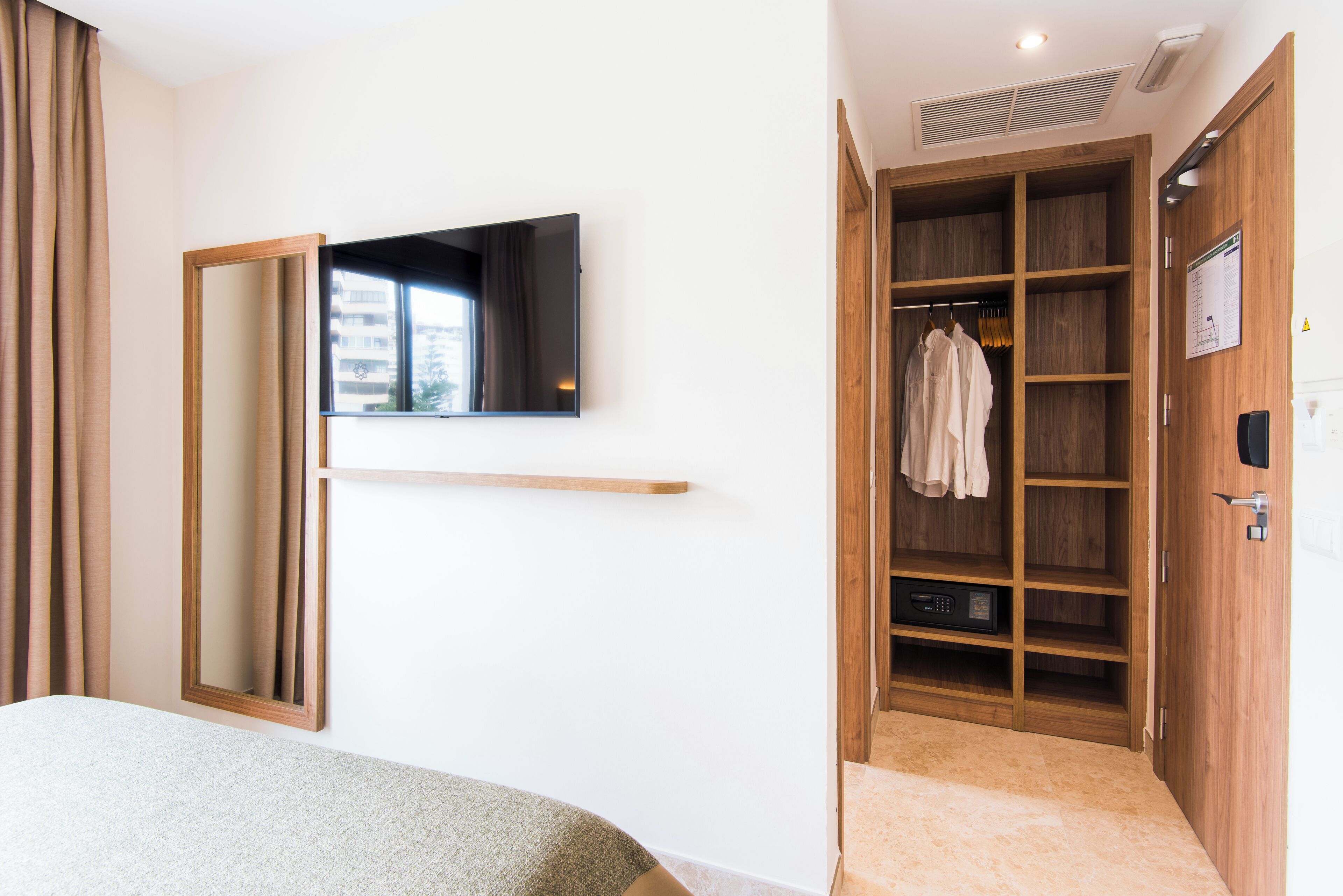 A hotel room with a bed, a TV mounted on the wall, a wooden wardrobe, and a mirror.