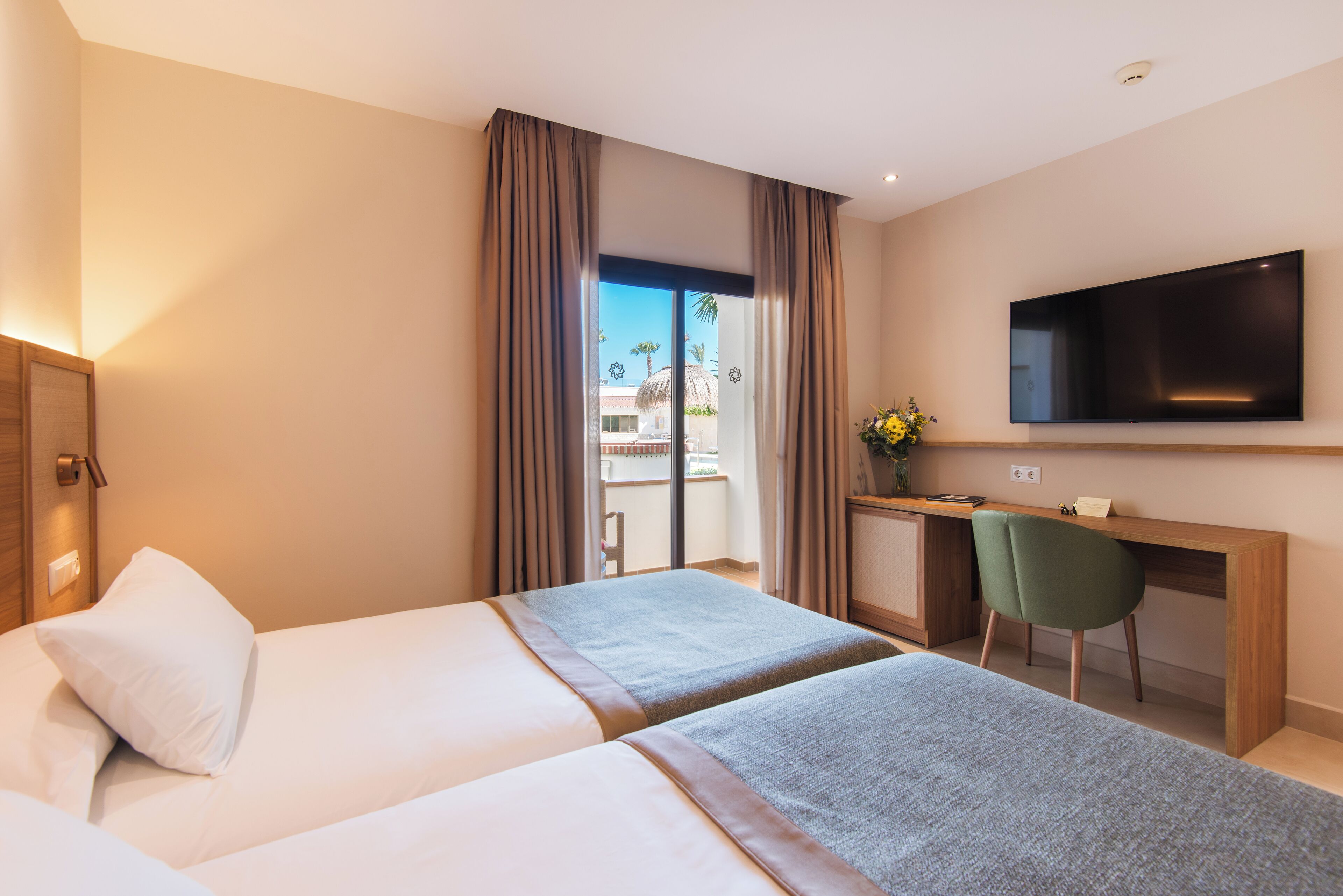 Une chambre d’hôtel avec deux lits, un bureau, une chaise, une télévision et une vue sur une piscine et des palmiers.