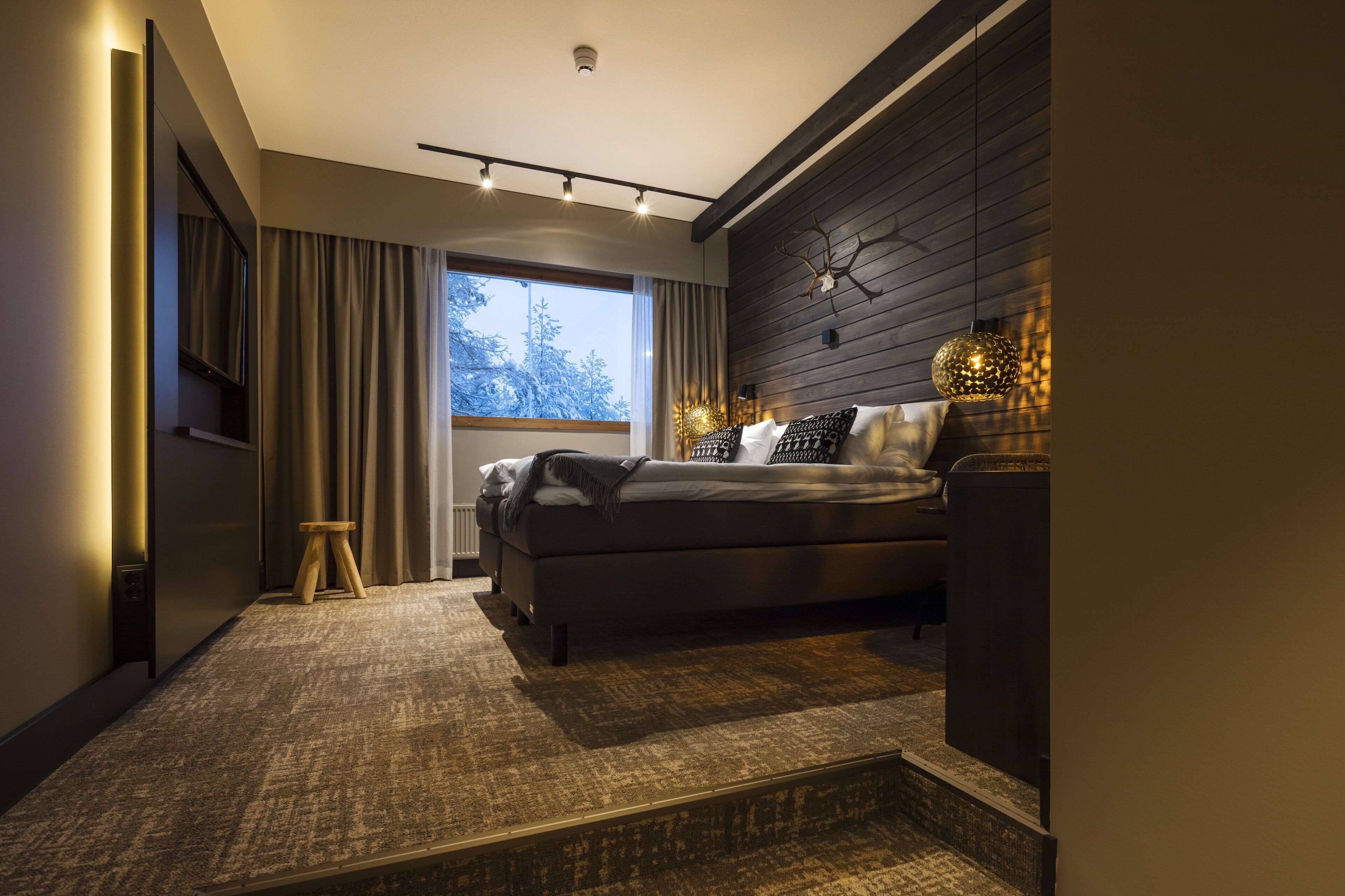 Un dormitorio moderno con una cama grande, un taburete pequeño, un televisor y una ventana con cortinas.