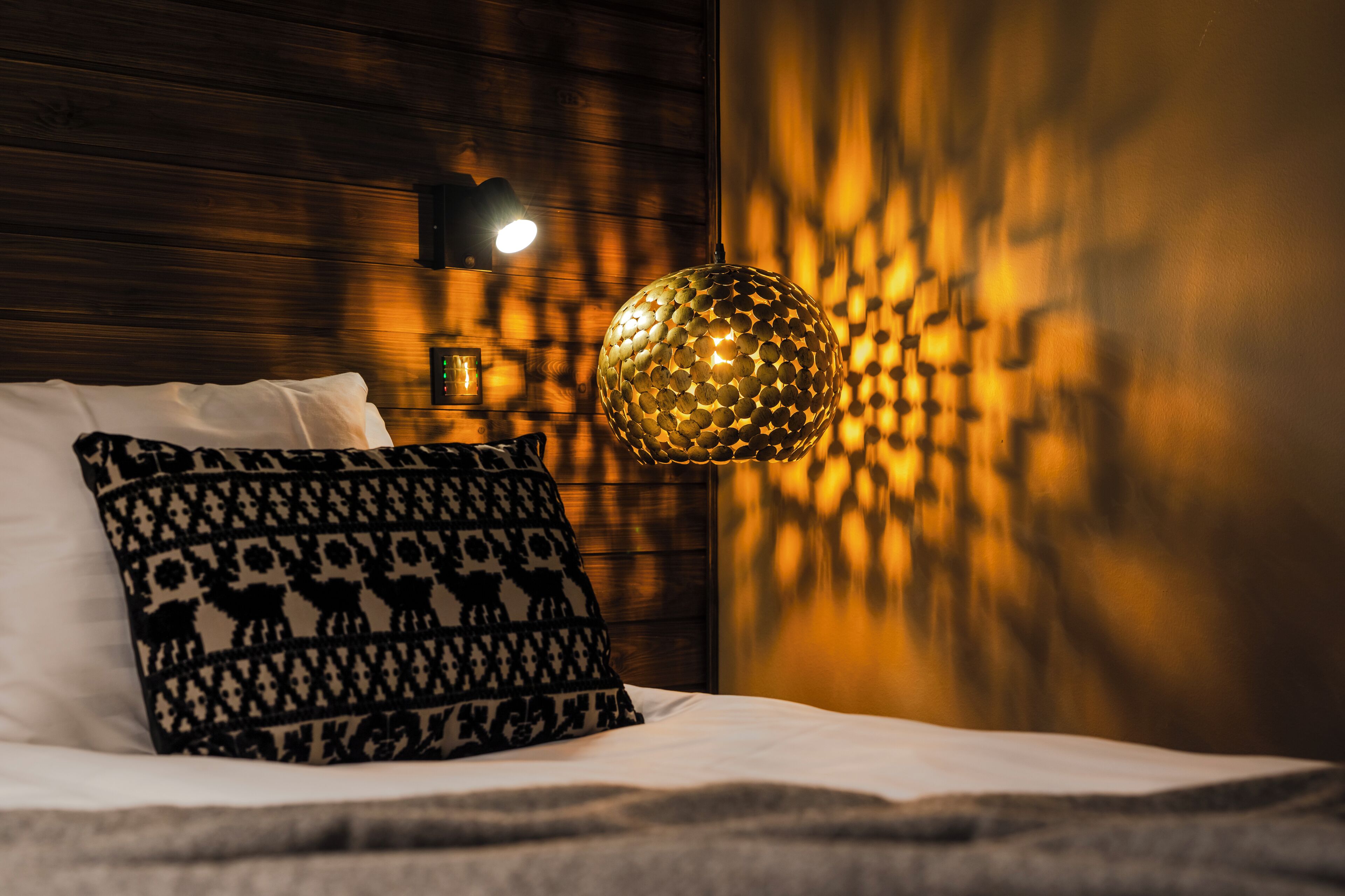 A neatly made bed with patterned pillows, a wall-mounted light fixture, and a textured wall.