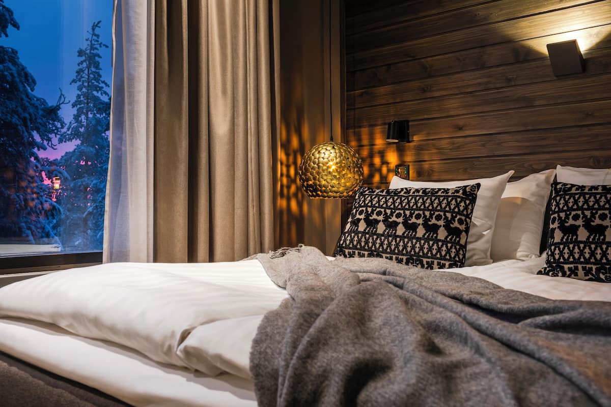 Un dormitorio con cama, cortinas, almohada estampada, lámpara fijada a la pared y vistas de árboles al exterior.