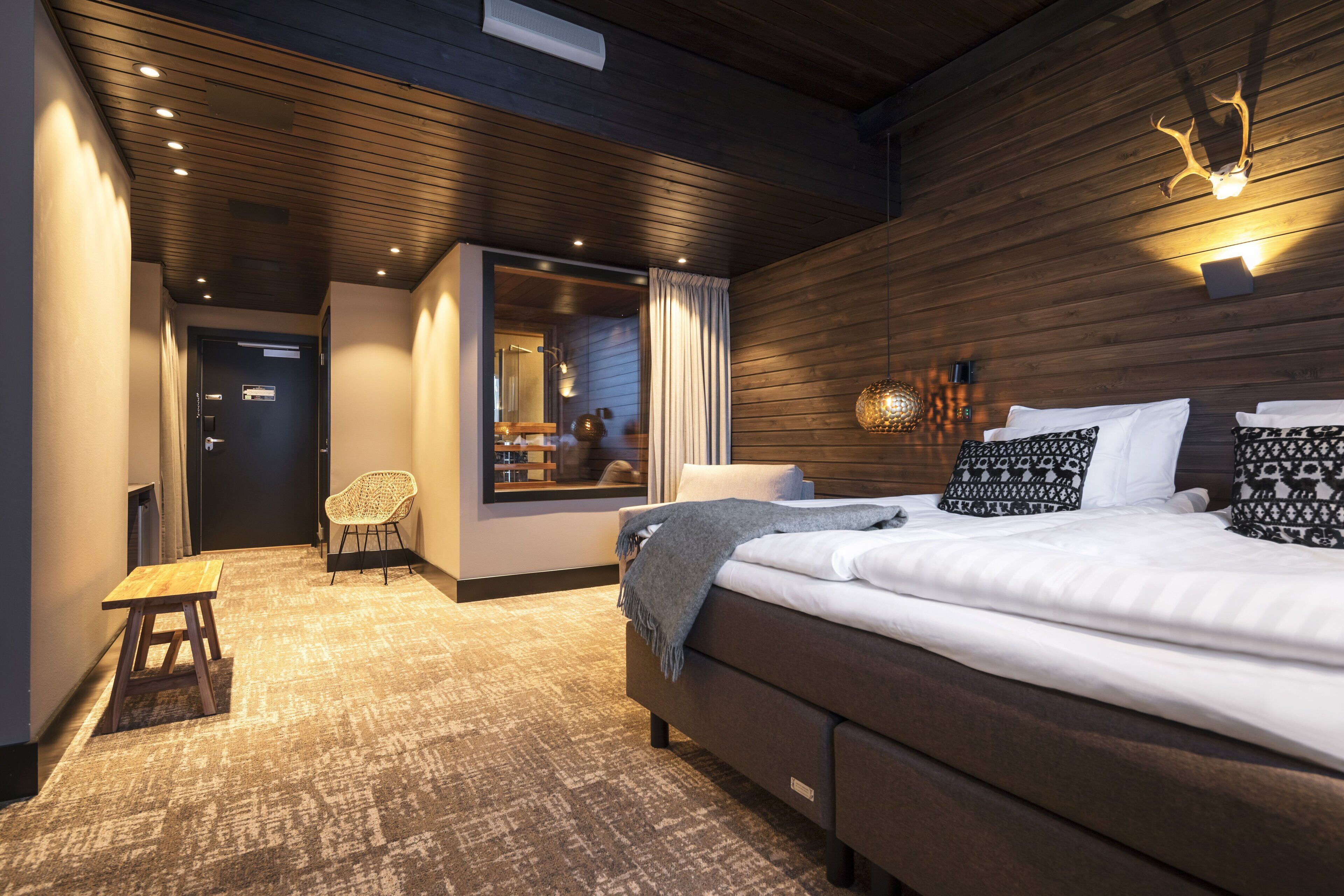 A modern hotel room with a large bed, wooden ceiling, and a view through a window.