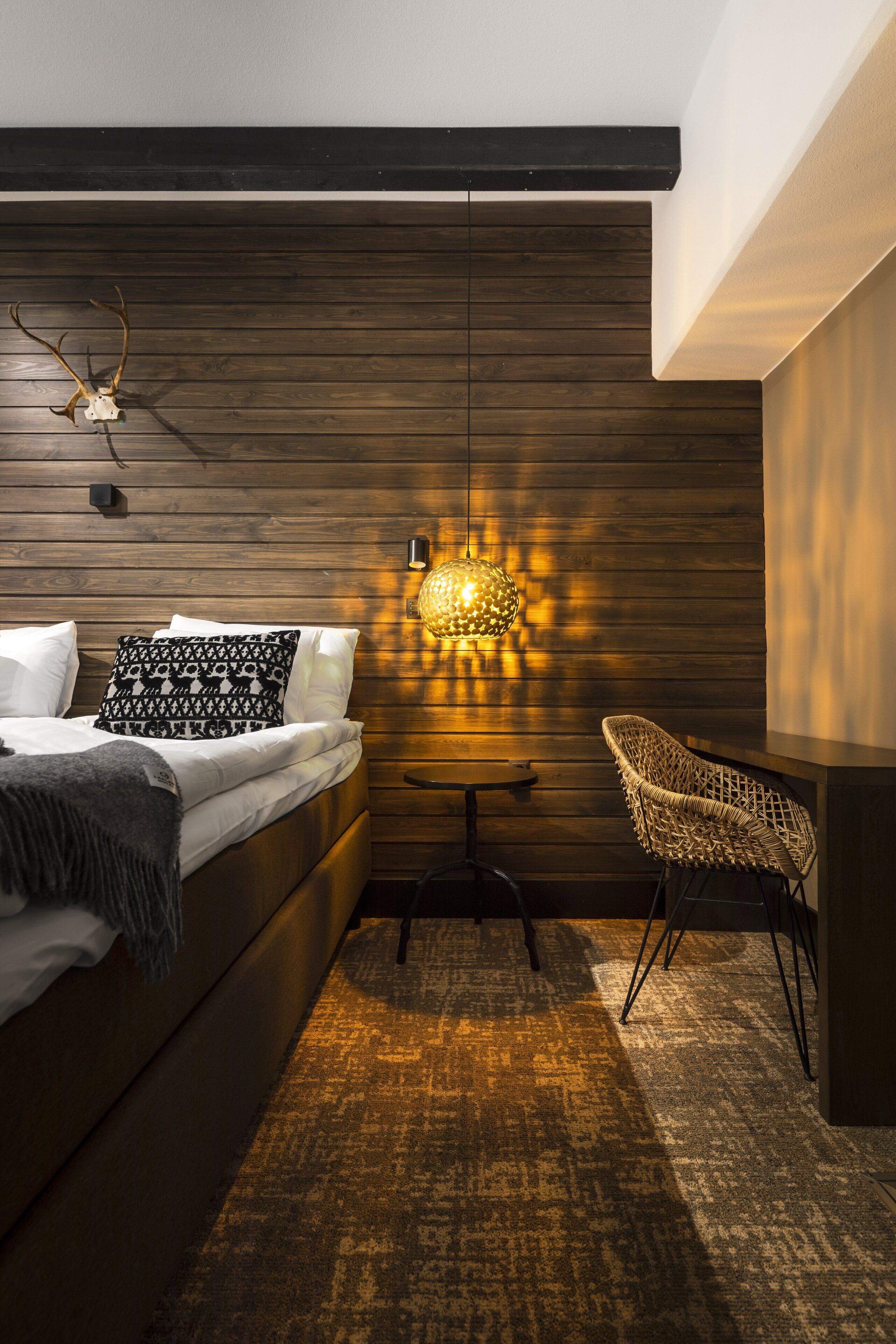 Un dormitorio moderno con un panel de madera en la pared, una cama con almohadas estampadas, un sillón tejido y una mesa con una lámpara original.