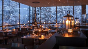 Ein Essbereich mit gedeckten Tischen, Kerzen und Blick auf verschneite Bäume durch große Fenster.