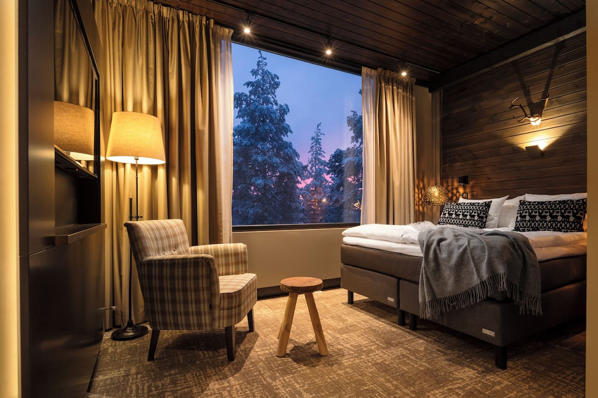 Un dormitorio con cama, una silla, una mesita, una lámpara y una ventana con cortinas.