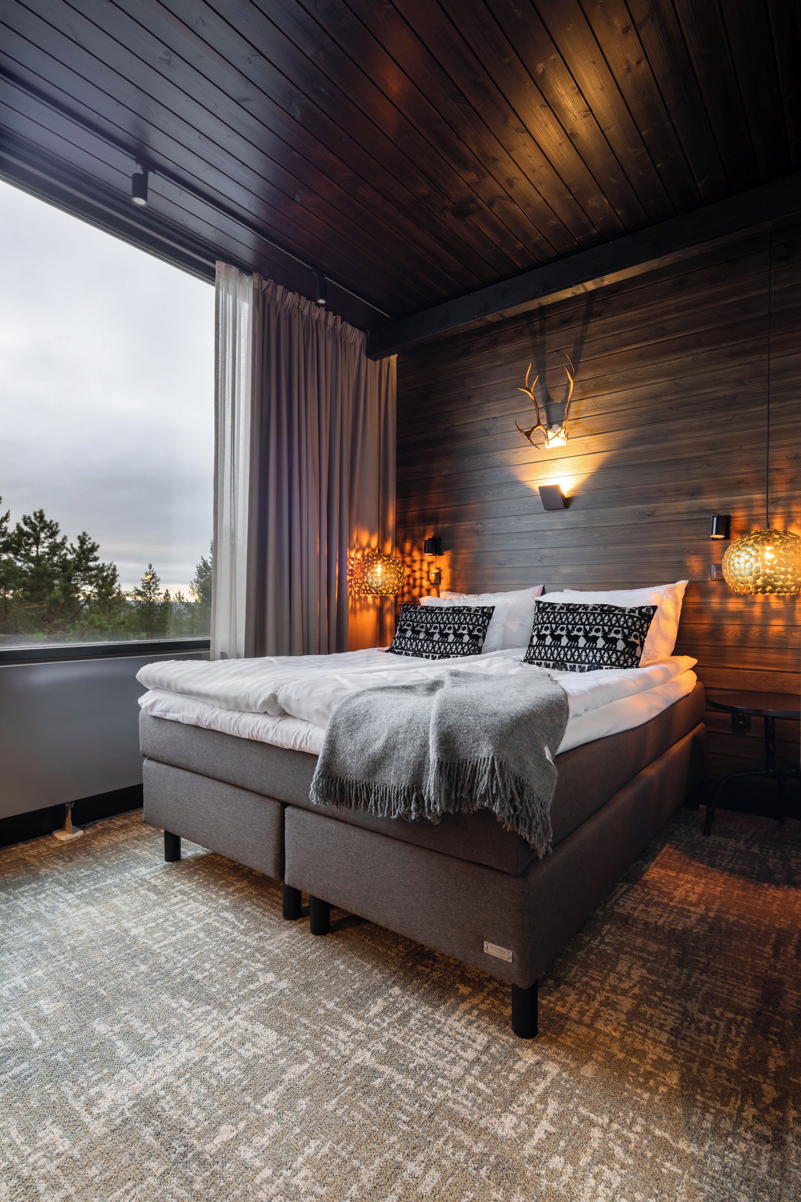 A modern bedroom with a large bed, a view of the outdoors, and a wooden ceiling.