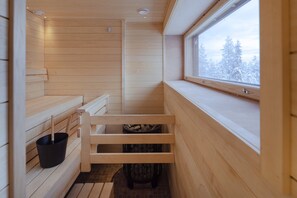 Una sala de sauna con bancos de madera, un cubo negro y una ventana con vistas nevadas.