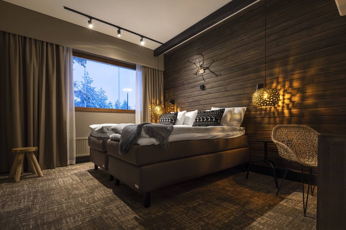 Un dormitorio moderno con una cama grande, un taburete pequeño, una silla de mimbre y una ventana con cortinas.