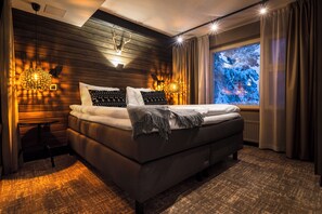Camera d'hotel moderna con un letto grande, pareti in legno, vista su alberi innevati e un tavolino con una lampada.