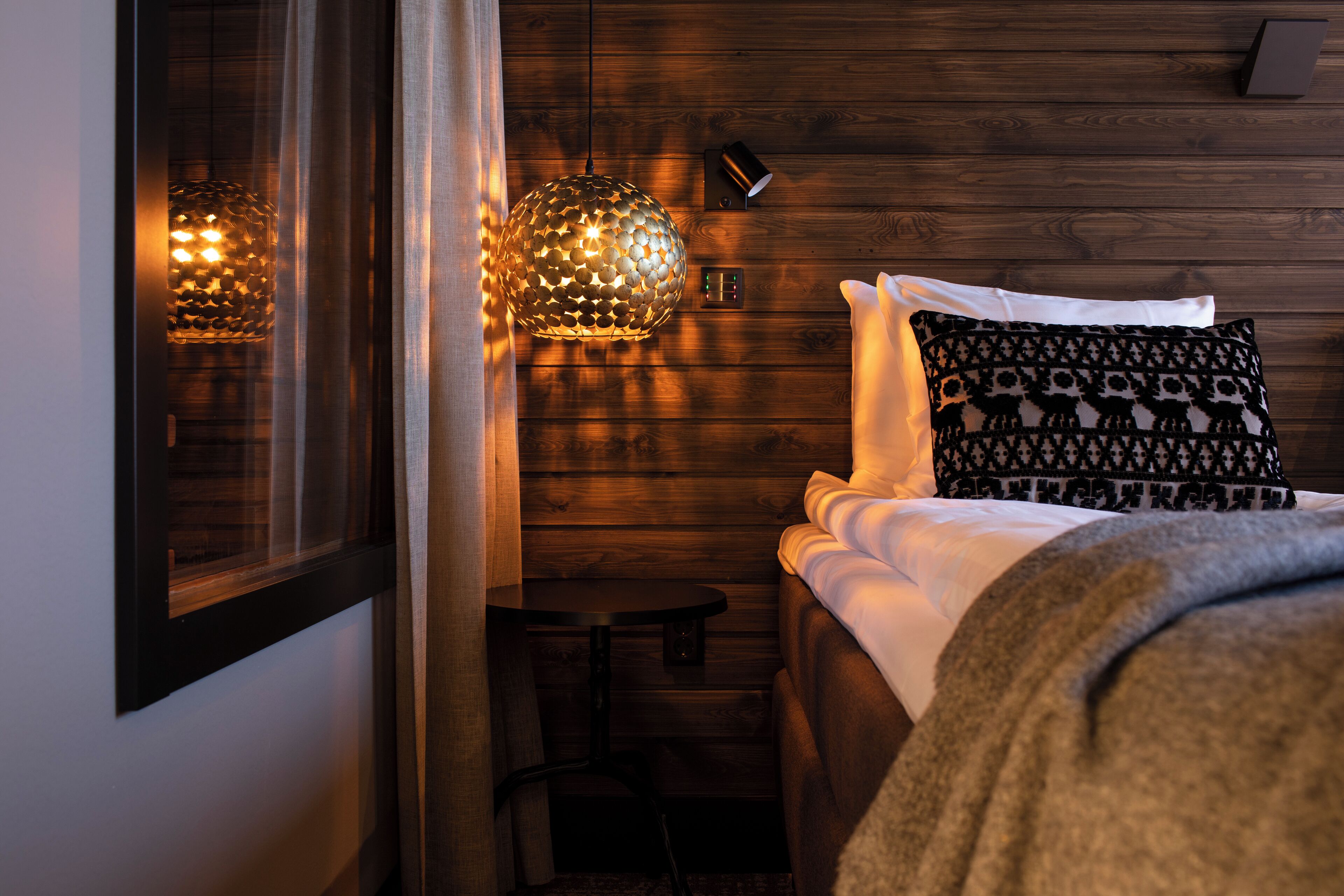 Un dormitorio acogedor con pared de madera, una cama con ropa de cama blanca, un cojín estampado y dos lámparas colgantes.