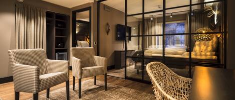 A modern hotel room with a large sliding glass door leading to a balcony, two checkered armchairs, a wicker chair, a flat-screen TV, and a bed with white linens.