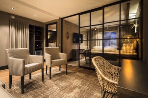 Una habitación de hotel moderna con un amplio ventanal corredero que da a un balcón, dos sillones a cuadros, una silla de mimbre, un televisor de pantalla plana y una cama con ropa blanca.
