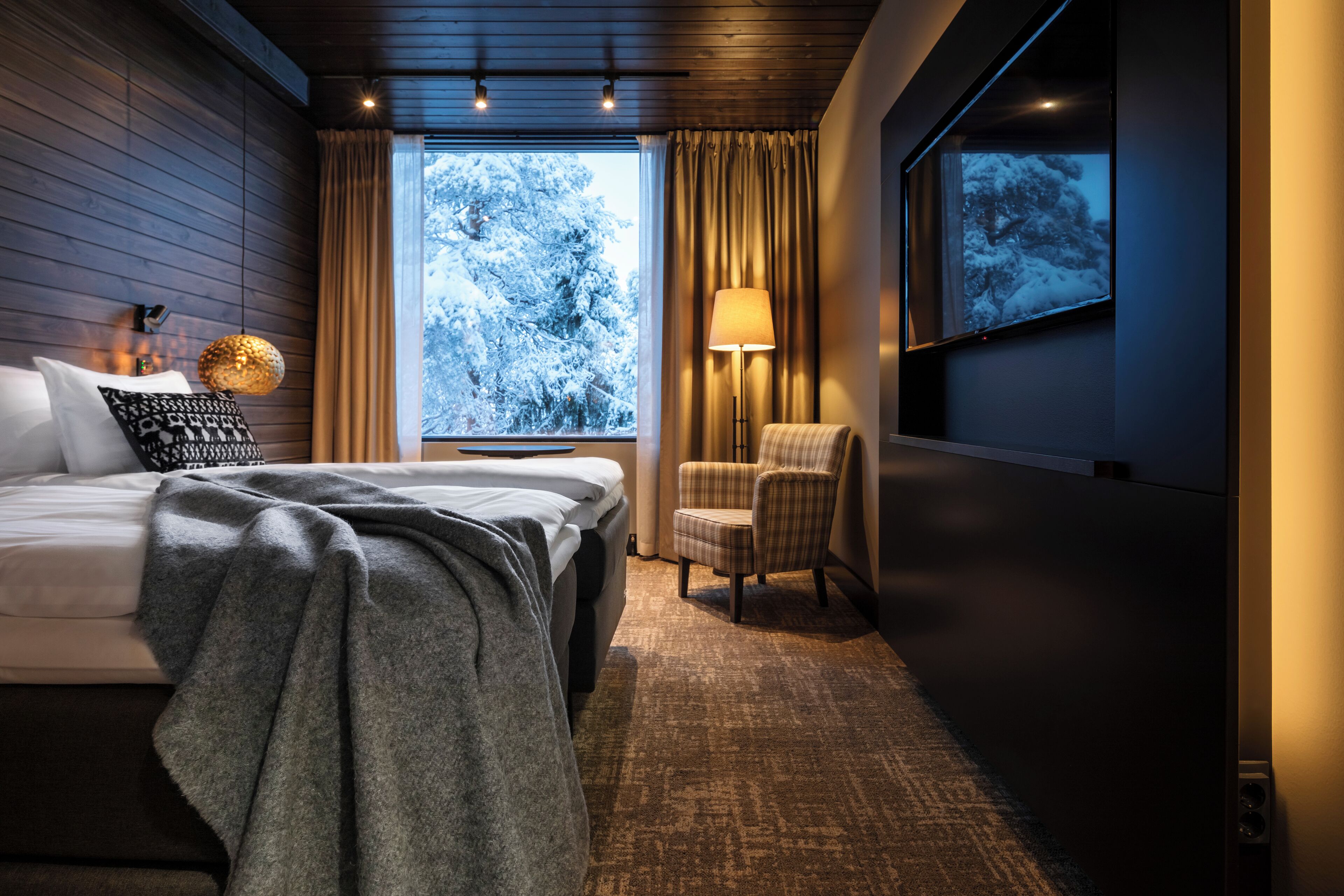 Habitación de hotel moderna con cama, silla, lámpara y ventana grande que ofrece vistas a árboles cubiertos de nieve.