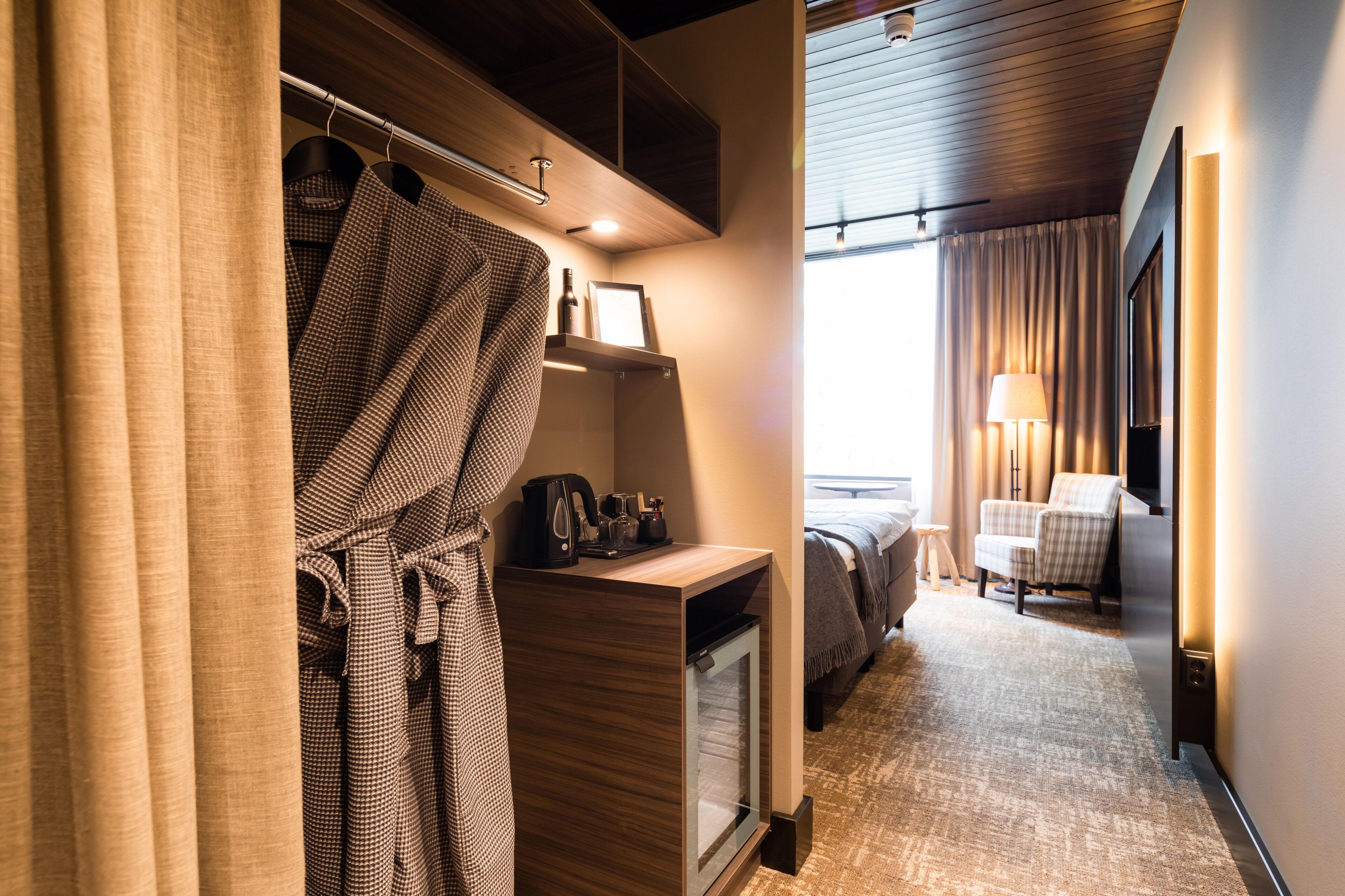 A hotel room with a wooden desk, a bed, a chair, and a robe hanging on a rack.