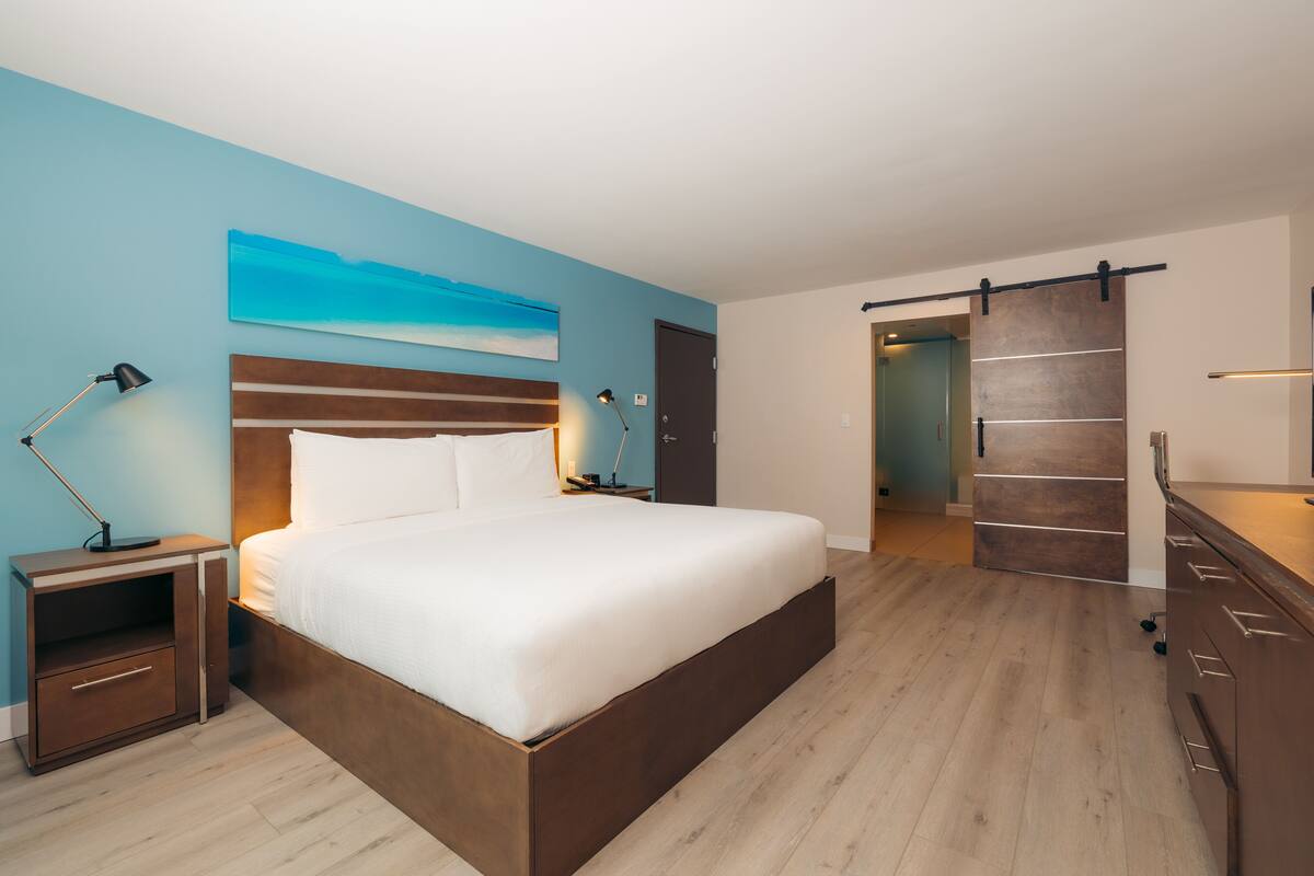 A modern bedroom with a large bed, a nightstand, a desk, and a sliding door leading to another room.