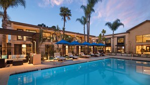 A pool area with lounge chairs and umbrellas, surrounded by palm trees and a building with multiple balconies. - The Cove Hotel, Long Beach (Long Beach)