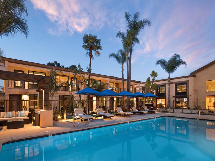 Zona de piscina con sillones de descanso y sombrillas, rodeada de palmeras y un edificio con varios balcones.