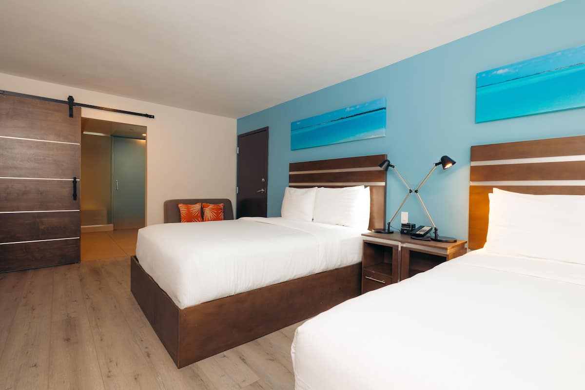 A hotel room with two beds, a wooden floor, a desk with a lamp, and artwork on the walls.