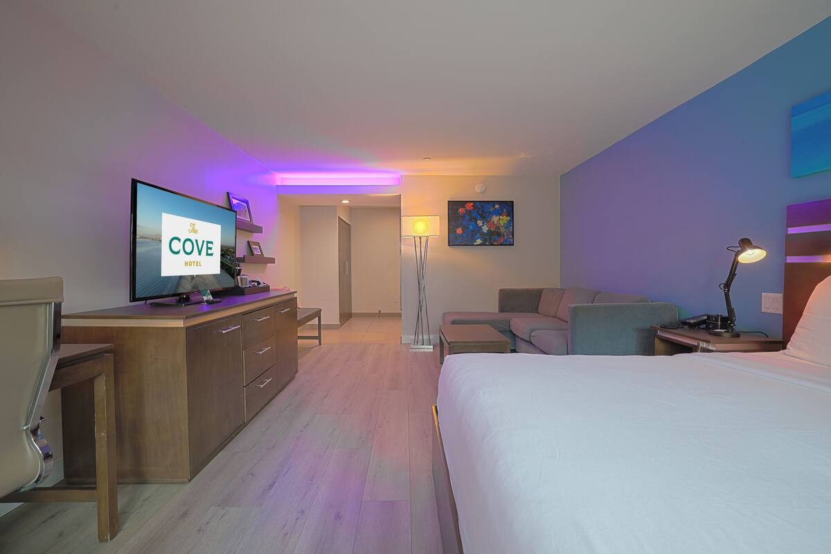 A hotel room with a bed, a desk with a computer, a TV displaying 'The Cove Hotel', a lamp, and a painting on the wall.