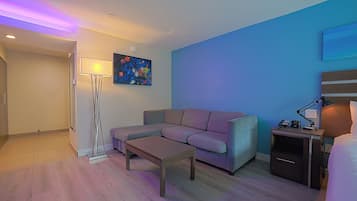 A modern hotel room with a sofa, a small table, a nightstand with a lamp, and a painting on the wall.
