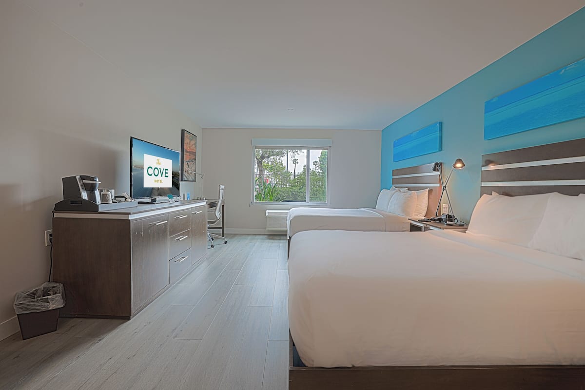 A hotel room with a bed, a desk, a coffee machine, a trash bin, and a window with a view of greenery.